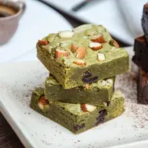 Fotografia de três pedaços de brownie de pistache empilhados, com castanhas por cima.