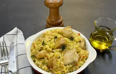 Foto da receita de arroz com peixe cremoso servida em uma panelinha de porcelana branca, ao lado esquerdo há talheres prateados sobre um pano quadriculado branco e preto. Ao fundo, um dispenser de azeite de vidro e um moedor de sal