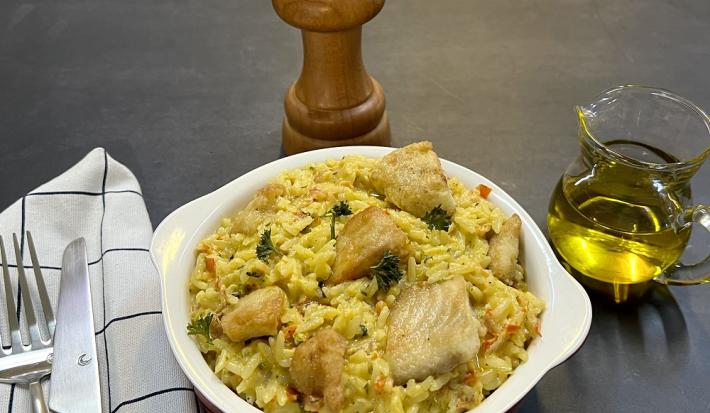 Foto da receita de arroz com peixe cremoso servida em uma panelinha de porcelana branca, ao lado esquerdo há talheres prateados sobre um pano quadriculado branco e preto. Ao fundo, um dispenser de azeite de vidro e um moedor de sal